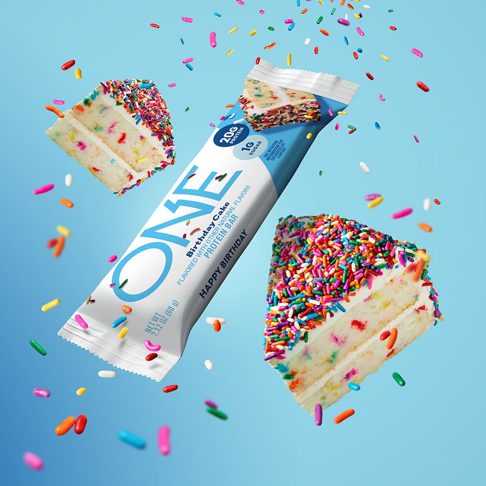 one bars birthday cake flavored protein bar beside slices of cake and sprinkles